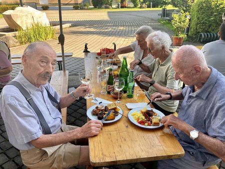 Bewohnerinnen und Bewohner sitzen an Tischen und genießen Essen und Getränke während des Grillfestes