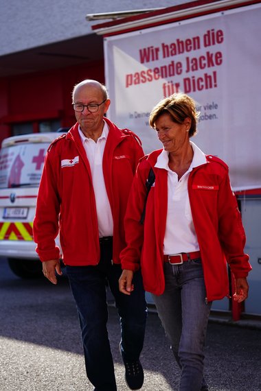 Zwei Rotkreuz Mitarbeiter:innen beide in roter Weste gehen zu ihrem Essen auf Rädern Auto am Anfang ihrer Schicht.