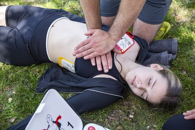 Ein Ersthelfer führt bei einer bewusstlosen Frau eine Herzdruckmassage durch; die Elektroden eines Defibrillators sind bereits angebracht.