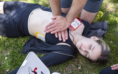 Ein Ersthelfer führt bei einer bewusstlosen Frau eine Herzdruckmassage durch; die Elektroden eines Defibrillators sind bereits angebracht.