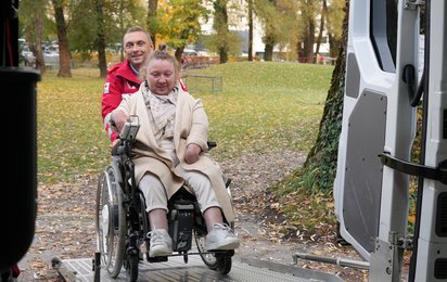 Eine im Rollstuhl sitzende Dame wird von einem Sanitäter in roter Uniform in einen Rettungswagen geschoben.