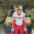 Katja und Achim befinden sich vor einem Rettungswagen. Er trägt sie dabei huckepack und beide lachen dabei.
