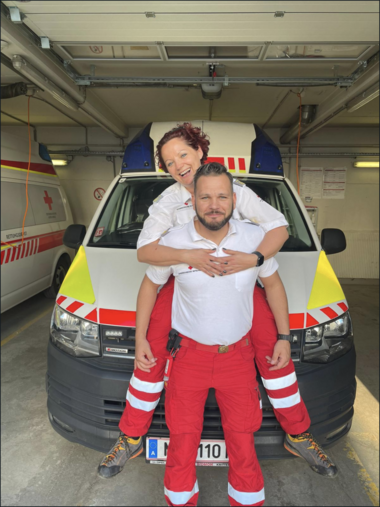 Katja und Achim befinden sich vor einem Rettungswagen. Er trägt sie dabei huckepack und beide lachen dabei.