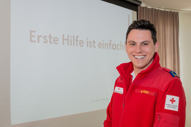 Weiss Moritz - Erste-Hilfe-Lehrbeauftragter - steht in einem Lehrsaal. Im Hintergrund sieht man den Spruch "Erste Hilfe ist einfach".