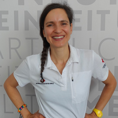 Dr. Andrea Schmid Schirmer - © Rotes Kreuz Tirol Christine Widmann Portrait von Andrea Schmid Schirmer im Roten Kreuz Poloshirt vor der Wand mit den Rotkreuz Grundsätzen