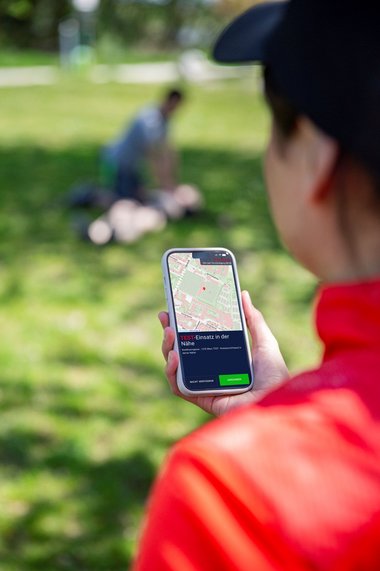Eine Person hält ein Smartphone in der Hand, auf dessen Anzeige eine Landkarte und der Text "Test-Einsatz in der Nähe" zu sehen sind.