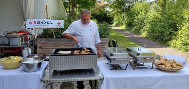 Ein Koch mit Grillzange an einem Griller, daneben am Tisch Schüsseln mit Essen, ein Brotkorb und zwei Warmhaltebehälter