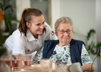 Eine Rotkreuz-Mitarbeiterin lehnt sich über eine Seniorin, sie lächeln in die Kamera.