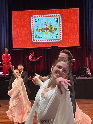 Eine Tänzerin in einem weißen Kleid streckt ihren Arm lächelnd in Richtung Kamera.