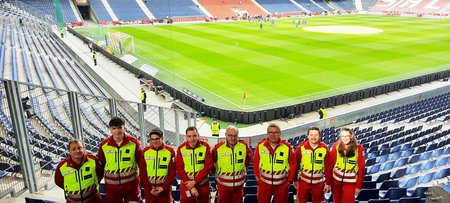 Gruppenaufnahme von acht Rettungssanitäterinnen. Sie stehen nebeneinander im leeren Stadion mit dem Rücken zu noch nicht bespielten Spielfeld.