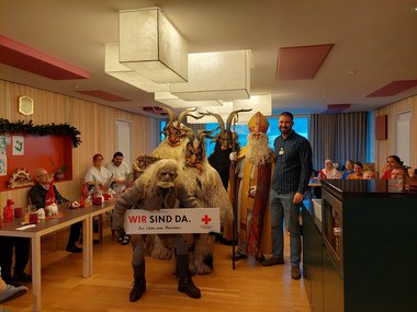 Nikolaus und vier Krampusse gemeinsam mit Lukas Utzmeier, Bewohnerinnen und Mitarbeiterinnen des Seniorenwohnhauses Hallein. Ein Krampus hält ein weißes Schild mit der Aufschrift "Wir sind da. Aus Liebe zum Menschen" sowie dem Logo des Roten Kreuzes.