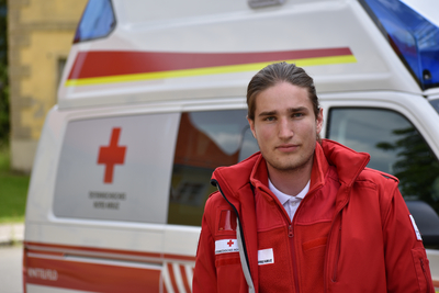 Sanitäter David steht vor einem Rettungswagen und trägt die markante rote Uniformjacke.