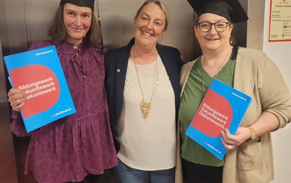 Drei Damen stehen vor einem Aufzug. Die Damen links und rechts halten eine Mappe mit einer Ausbildungsbescheinigung in der Hand und tragen einen Doktorhut. Die rechte Dame ist Conny Seeburger.