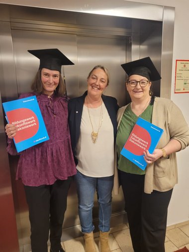 Drei Damen stehen vor einem Aufzug. Die Damen links und rechts halten eine Mappe mit einer Ausbildungsbescheinigung in der Hand und tragen einen Doktorhut. Die rechte Dame ist Conny Seeburger.