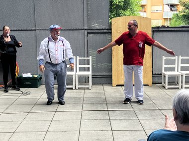 Der Clown steht links neben einem Pflege-Mitarbeiter, der die Arme seitlich wegstreckt