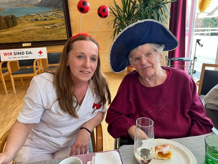 Eine Bewohnerin mit Hut sitzt gemeinsam mit einer Pflegemitarbeiterin mit einem Haarreifen mit Fußbällen an einem Tisch. Vor ihr liegt ein halb gegessener Faschingskrapfen am Teller .
