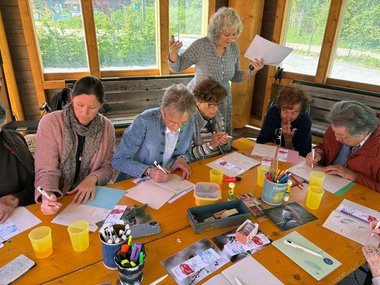 Eine Gruppe von Menschen schreibt auf Zetteln unter Anleitung einer Dame.