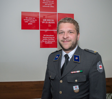 Mag. Daniel Miedl - Bezirksfreiwilligenkoordinator - steht in Ausgangsuniform vor einem Roten Kreuz, in dem einige Sätze des Leitbildes gedruckt sind.