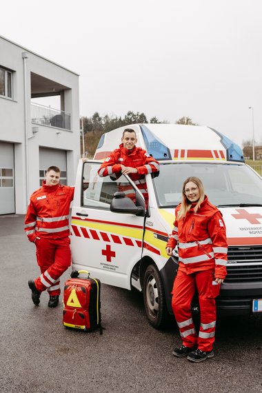 Drei junge Sanitäter:innen versammelt um ein Rettungsauto.
