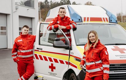 Drei junge Sanitäter:innen versammelt um ein Rettungsauto.