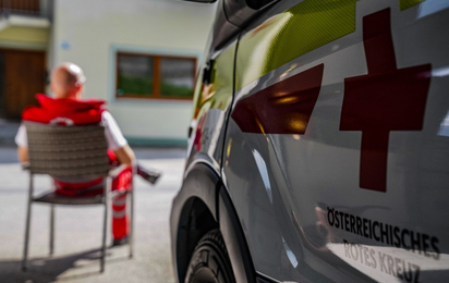 © Rotes Kreuz Schwaz / Hochschwarzer Sanitäter sitzt vor der Dienststelle, Rettungstransportwagen mit Logo auf rechter Seite