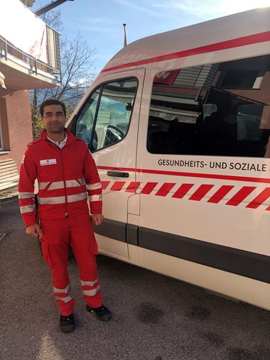 Am Bild zu sehen ist Muhammad neben dem Rettungswagen. Muhammad ist beim Roten Kreuz für den betreuten Fahrdienst zuständig und bringt KlientInnen an ihren Bestimmungsort