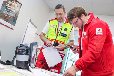 Links zu sehen ist Doktor Alexander Franz in einer Notarzt-Uniform mit Gilet, rechts ein Notarzt in Rotkreuz-Uniform. Vor ihnen steht auf einem Tisch ein Corpuls-Gerät zur Überwachung von Vitalparametern von Patientinnen.