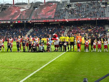 Blick auf das Spielfeld vor dem Anstoß. Spieler des FC Red Bull Salzburg und des SCR Altach haben links und rechts der Mittellinie Aufstellung genommen. Zwischen ihnen stehen der Schiedsrichter und zwei Linienrichter sowie das Maskottchen des FC Red Bull Salzburg. Vor den Spielern und Schiedsrichtern stehen Kinder in weiß-rotem bzw. blauen Trikots des FC Red Bull Salzburgs.