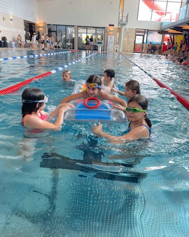 Ein Kind liegt in einem Schwimmbecken auf einer Luftmatraze, während es von fünf anderen Kindern im Wasser gezogen wird