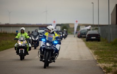 Ein Rotkreuz-Motorrad und ein Polizei-Motorrad fahren auf das Gelände zu - dahinter sieht man die anderen Motorräder.