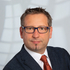 Portrait eines Mannes mit kurzen Haare, dunkler Brille und dunkelgrauem Sakko.