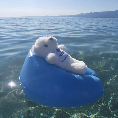 Erklärbär Bärli sitzt auf einem Wasserball treibend im Meer und genießt seinen Urlaub - natürlich hat er sich vorher mit Sonnencreme eingeschmiert.