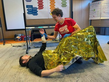 Ein Erste-Hilfe-Lehrbeauftragter in rotem Jugendrotkreuz-T-Shirt legt eine goldene Rettungsdecke über einen am Boden liegenden Teilnehmer des Kurses und hält dessen verbundenen linken Oberarm hoch.