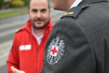 Zu sehen ist das Rotkreuz-Wappen auf dem Österreichischen Bundesadler, die den Ärmel eines Mitarbeiters in Ausgangsuniform ziert. Im Hintergrund sieht man (unscharf) einen Mitarbeiter in Rettungsdienst-Uniform.