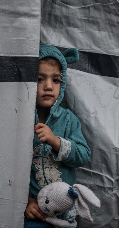 © ICRC / Abed Zaqout Ein kleines Mädchen in Gaza schaut vorsichtig hinter einem Zelt hervor und hält ihren schmutzigen Stoffhasen in der Hand.