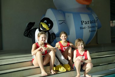 Drei Kinder sitzen auf Stufen, dahinter sitzt ein Pinguin-Maskottchen. Im Hintergrund ist eine große, aufgeblasene Bade-Ente mit weiß-blauen Streifen zu sehen.