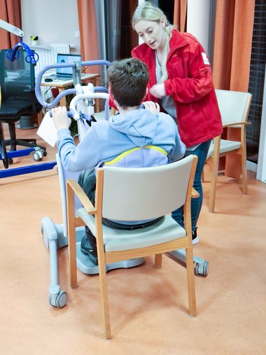Eine Frau in roter Jacke spricht mit einem jungen Mann auf einem Sessel, vor dem Hebekran steht.