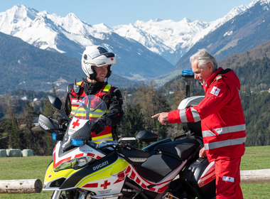 Motorradstaffel Rotes Kreuz - © Rotes Kreuz Tirol Daniel Liebl Rotkreuz-Präsident Günther Ennemoser in Roter Uniform und Jan Reiter in der Ausstattung der Motorradfahrer posieren gemeinsam mit dem RotKreuz-Motorrad vor der Tiroler Bergkulisse