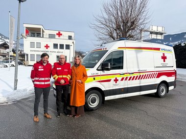 Drei Personen stehen vor einem Rettungswagen des Roten Kreuzes. Im Hintergrund ist die Rotkreuz-Bezirksstelle Sankt Johann zu sehen. Von links nach rechts: Daniel Kofler (Bezirksgeschäftsführer Rotes Kreuz St. Johann), Peter Riepler (Bezirksrettungskommandant St. Johann), Eveline Huber (Bürgermeisterin St. Johann)