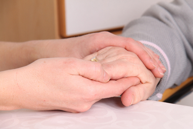 Die Hände einer jüngeren Frau umschließen eine Hand eines alten Menschen mit goldenem Ring und grauem Pulloverärmel.