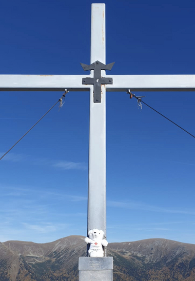 Zu sehen ist Bärli mit einem Gipfelkreuz und strahlend blauem Himmel dahinter.