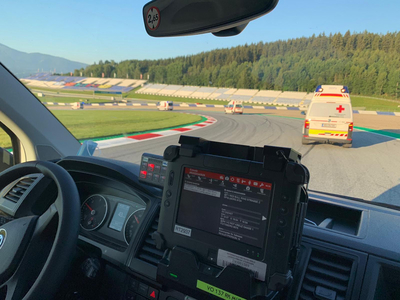 Einige Rettungswagen befinden sich auf einer Rennstrecke (dem Red Bull Ring) und beziehen grade ihre zugewiesenen Posten. Man sieht eine Fahrerkabine von Innen und weitere Rettungswagen vor dem Fahrzeug.