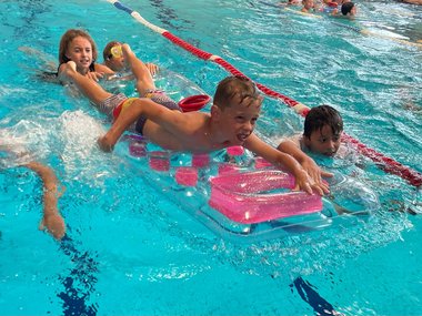 Ein Schüler liegt auf einer Luftmatratze im Wasser und wird von anderen Schüler:innen am Wasser bewegt.