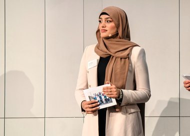 Junge Frau mit hellbeiger Jacke und braunem Kopftuch hält ein Heft in den Händen vor einer hellen Wand