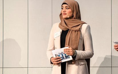Junge Frau mit hellbeiger Jacke und braunem Kopftuch hält ein Heft in den Händen vor einer hellen Wand