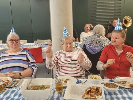Drei Bewohnerinnen sitzen mit weiß-blauen Partyhüten an einem Tisch mit weiß-blauer Tischdecke. Auf dem Tisch stehen für ein Oktoberfest typische Speisen und Getränke, wie z.B. Brezeln und Weißwürste.