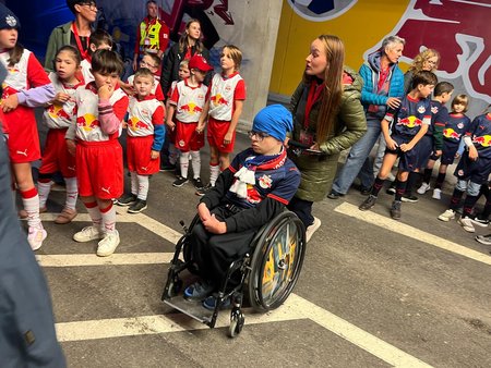Im Mittelpunkt ist ein in einem Rollstuhl sitzender Bub mit einem blauen FC-Red-Bull-Salzburg-Trikot und einen Schal um den Hals zu sehen. Hinter ihm steht eine Frau und hält den Bügel des Rollstuhls. Um die beiden herum stehen mehrere Kinder in weiß-roten bzw. blauen FC-Red-Bull_Salzburg-Trikots.
