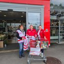 TÖT Aktionstag 2026 - © Österreichisches Rotes Kreuz / LV Niederösterreich Drei Personen mit Team-Österreich-Westen und -Jacke stehen vor einem Supermarkteingang neben einem Einkaufswagen mit haltbaren Lebensmitteln und einem Plakat zur Tafel-Aktion am 12.06.2026