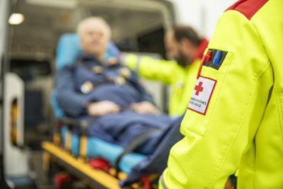 Eine Person in Einsatzkleidung des Roten Kreuzes steht vor einem Rettungswagen, während im Hintergrund ein Patient auf einer Trage versorgt wird. Die Szene zeigt einen Rettungseinsatz.