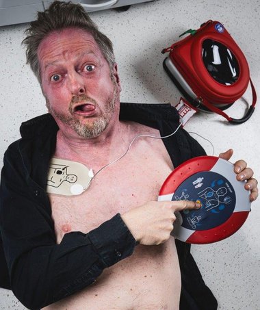 Markus Koschuh mit halbnacktem Oberkörper, liegt am Boden, die Defi-Paddles aufgeklebt